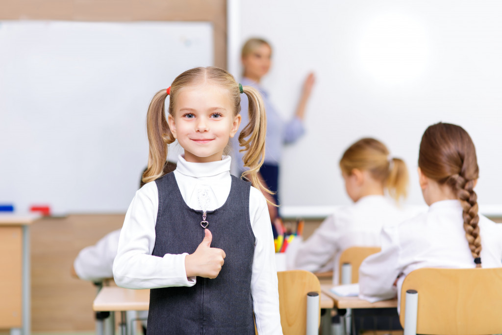 Like my school. Cute little girl is smiling lovely and showing thumbs up while standing in the classroom.