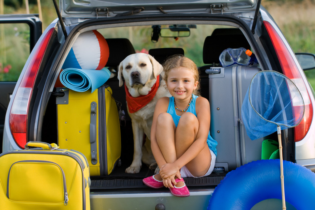young girl smiling while sitting at the back of her family's van next to dog and suitcases