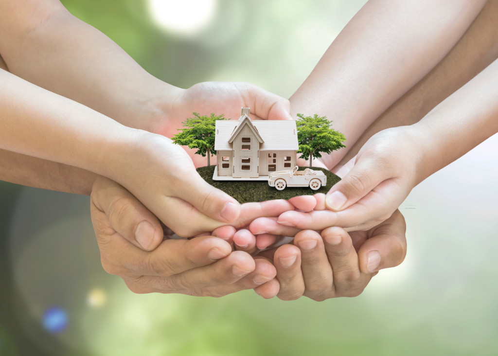 multiple hands holding a miniature house model with car and trees to represent community